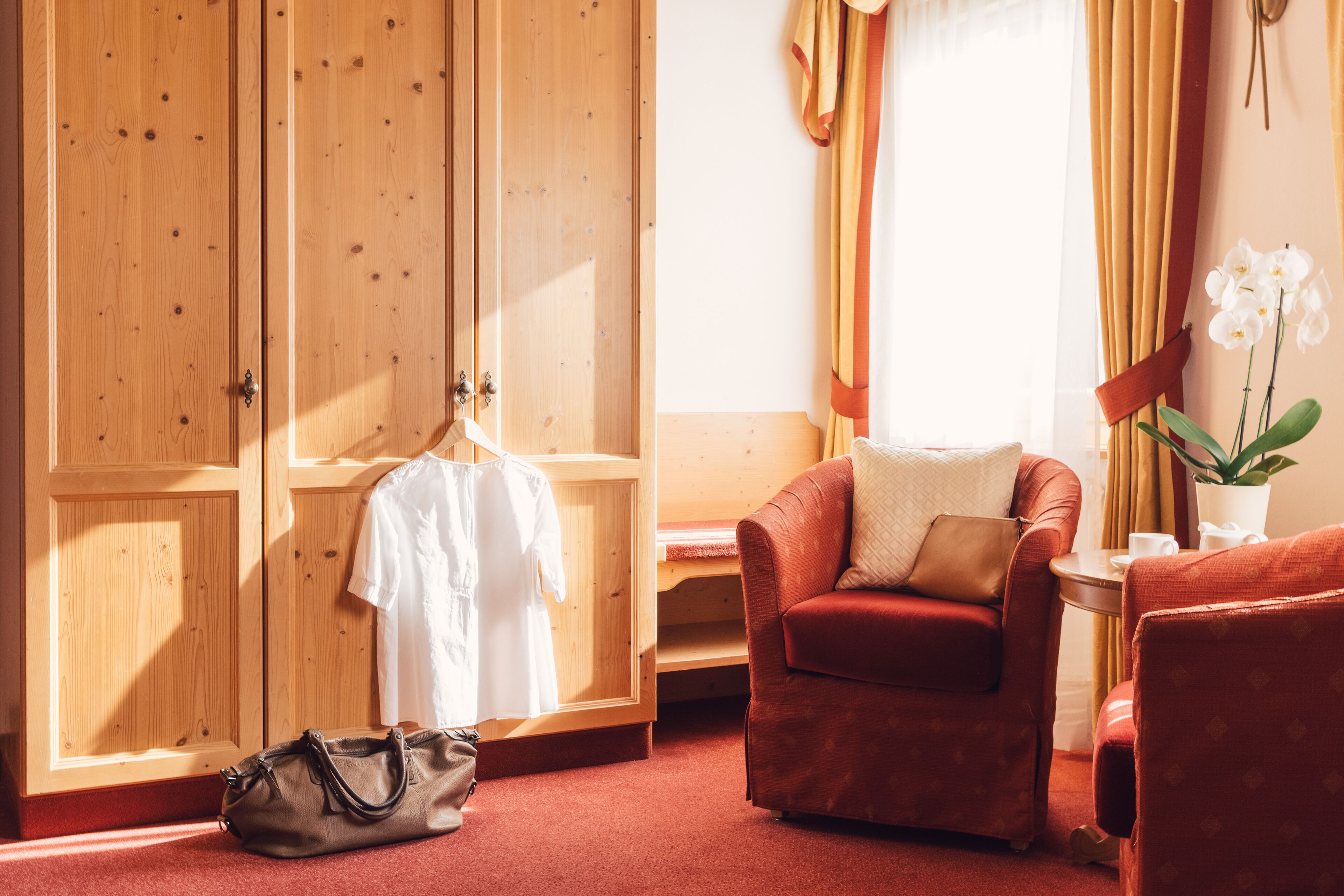 A hotel room with a wooden wardrobe, a red armchair, a small table with a potted orchid, and a hanging white shirt.