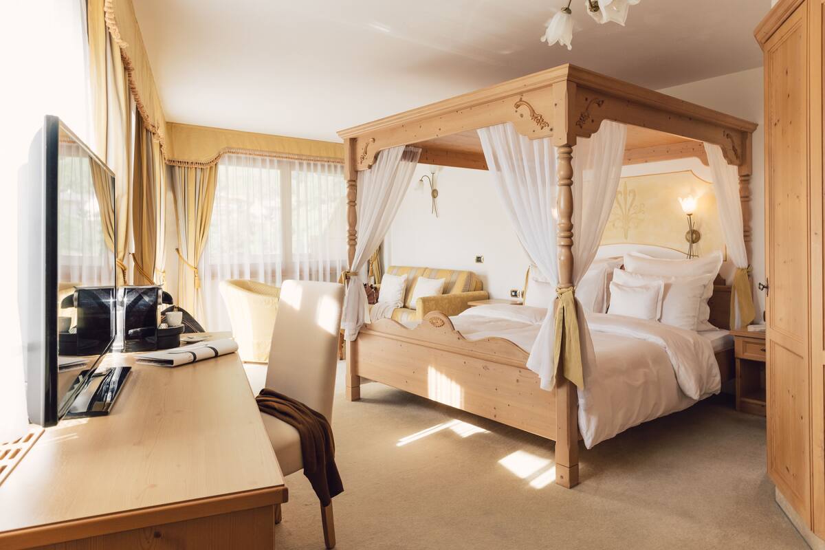 Een hemelbed met witte lakens, een houten bureau met stoel, en een nachtkastje met een lamp.