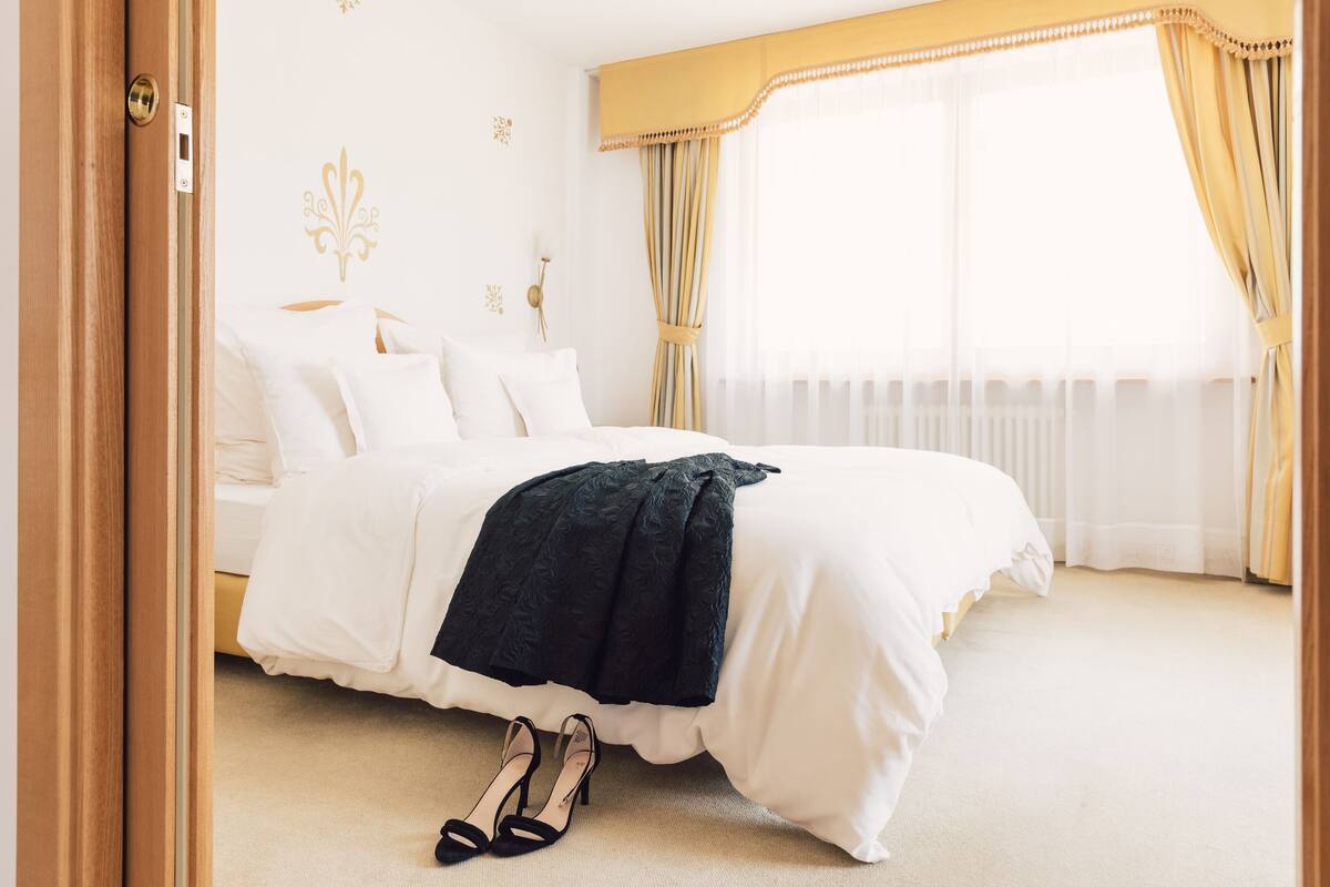 Een netjes opgemaakt bed met witte lakens en een donkere plaid, een paar zwarte hakken op de vloer, en een raam met dunne gordijnen.