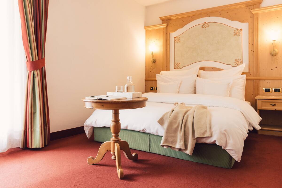 Een hotelkamer met een bed, een ronde houten tafel met een boek en glazen, een groen gestoffeerd hoofdeinde en een nachtkastje met een lamp.