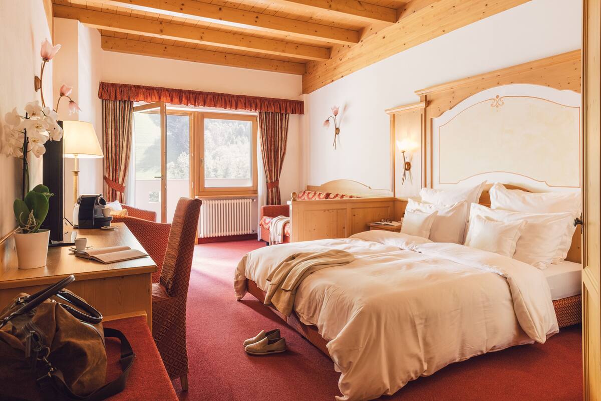 Een hotelkamer met een houten plafond, een bed met witte lakens, een bureau met een lamp, een stoel en een raam met gordijnen.