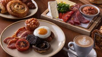 A breakfast spread with croissants, cheese, meats, beans, tomatoes, mushrooms, and a cup of coffee.