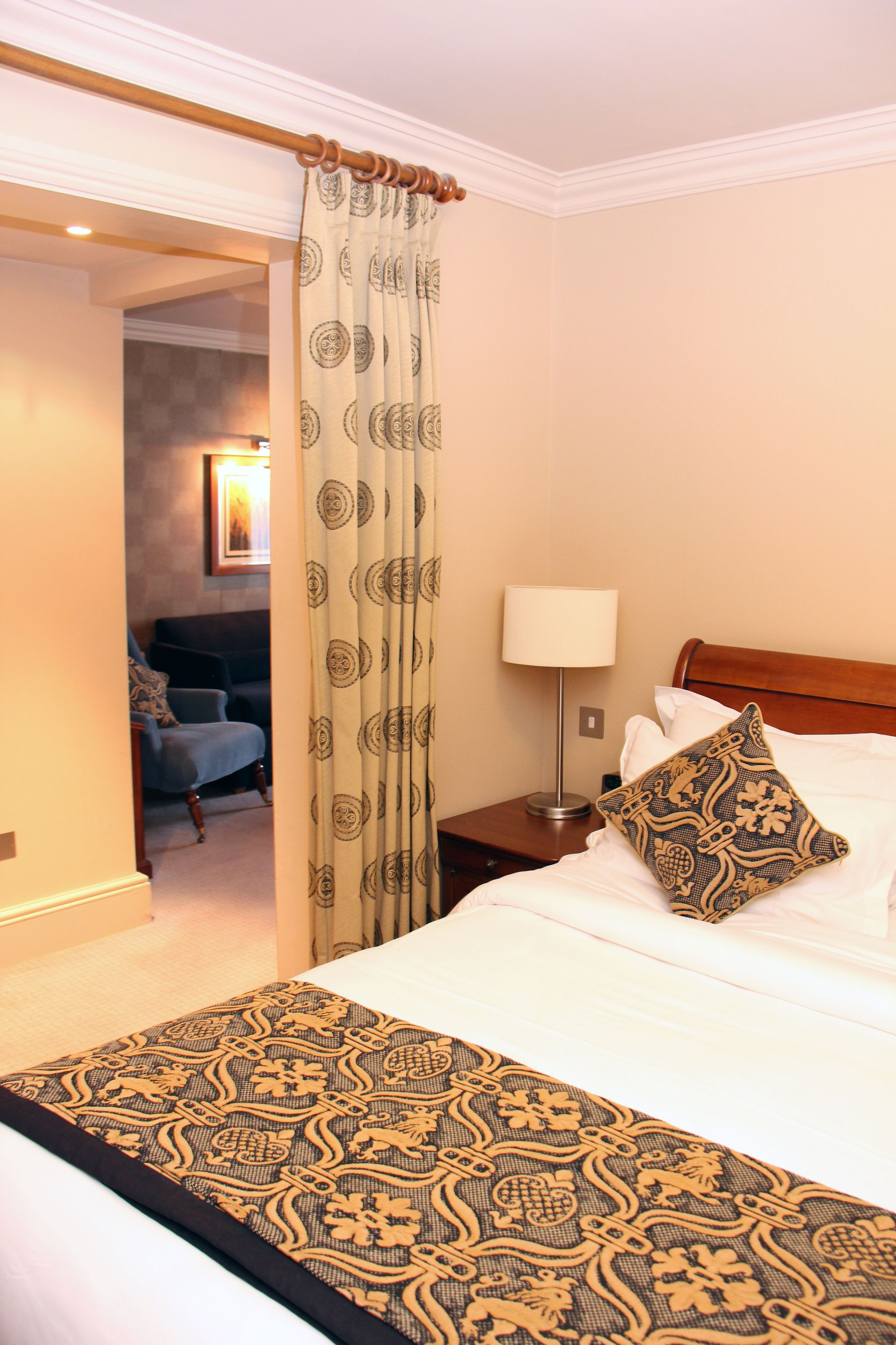 Habitación de hotel con cama, mesita de noche, lámpara, manta estampada y cortina.
