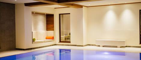 A modern indoor pool area with a clear pool, wooden beams on the ceiling, and a bench along the poolside.