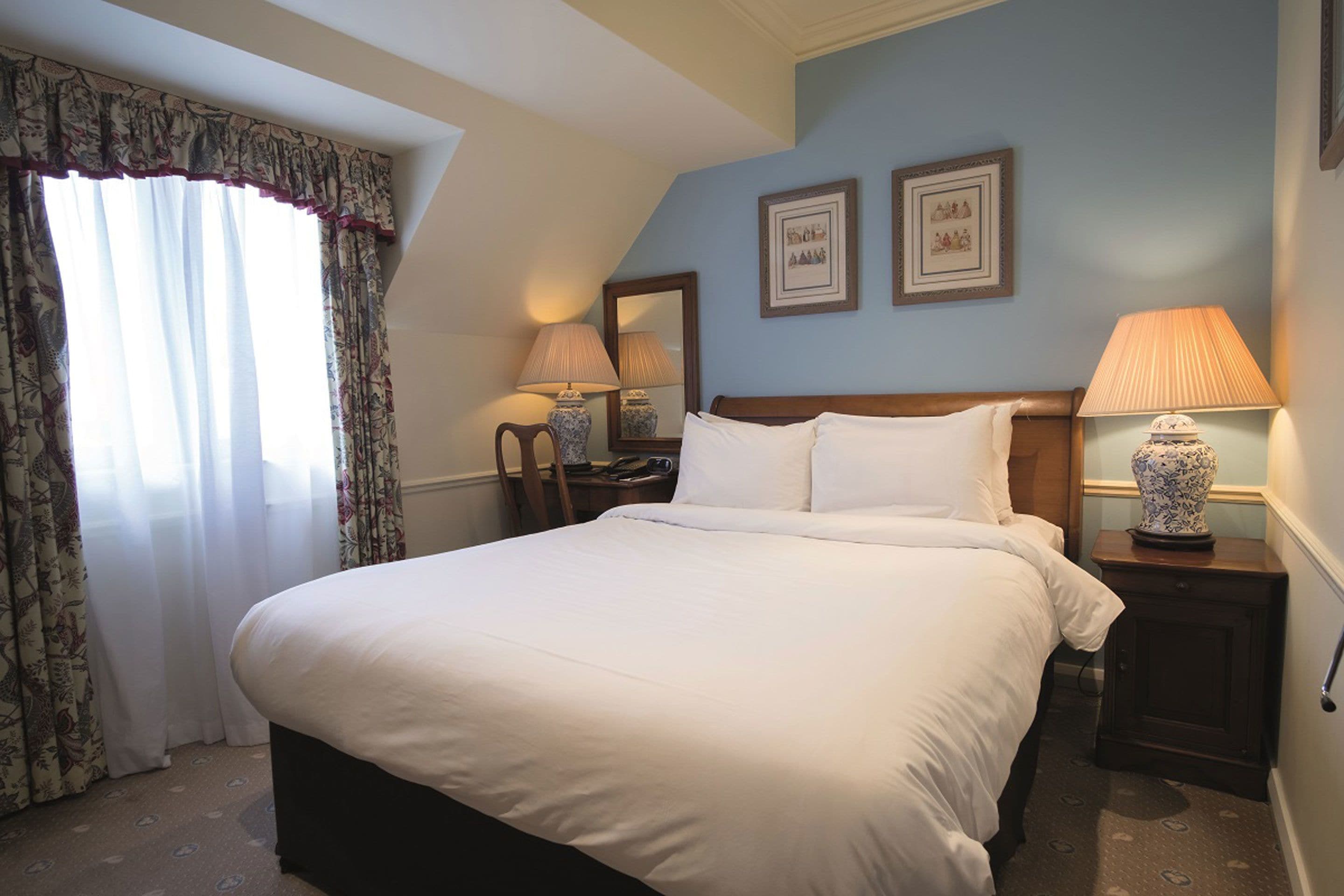A neatly made bed with white linens in a room with a sloped ceiling. There are two bedside tables with lamps, a large window with curtains, and two framed pictures on the wall.