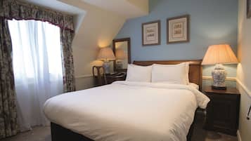 A neatly made bed with white linens in a room with a sloped ceiling. There are two bedside tables with lamps, a large window with curtains, and two framed pictures on the wall.