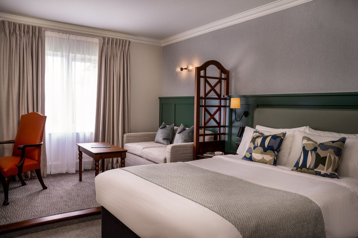 Una habitación de hotel con cama, una silla, una mesita, una lámpara y un aplique de pared.