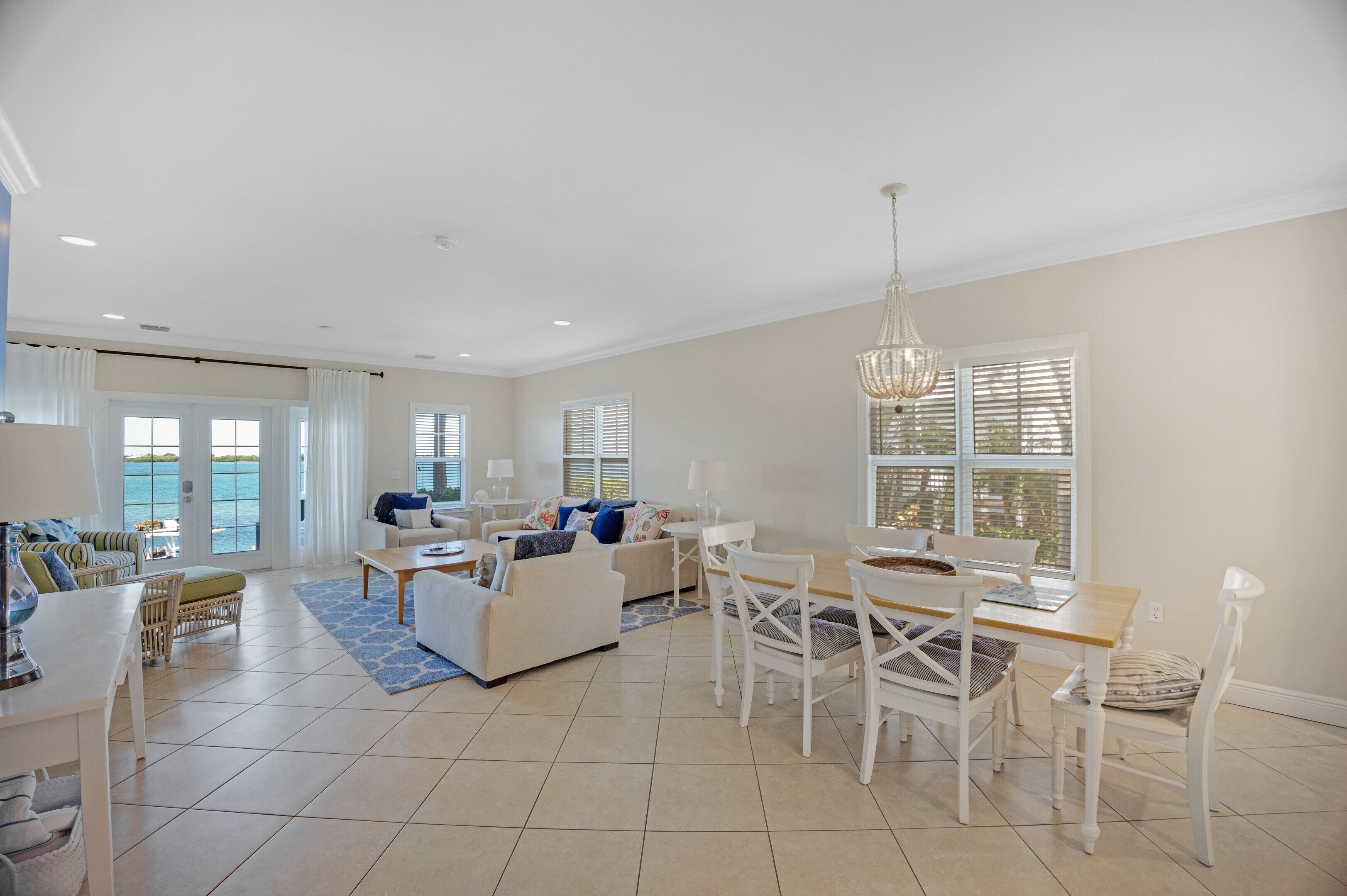 A spacious living area with a dining table, chairs, and a sofa. There are large windows with blinds, a chandelier, and a view of the ocean.