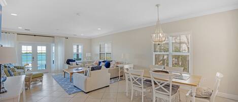 A spacious living area with a dining table, chairs, and a sofa. There are large windows with blinds, a chandelier, and a view of the ocean.