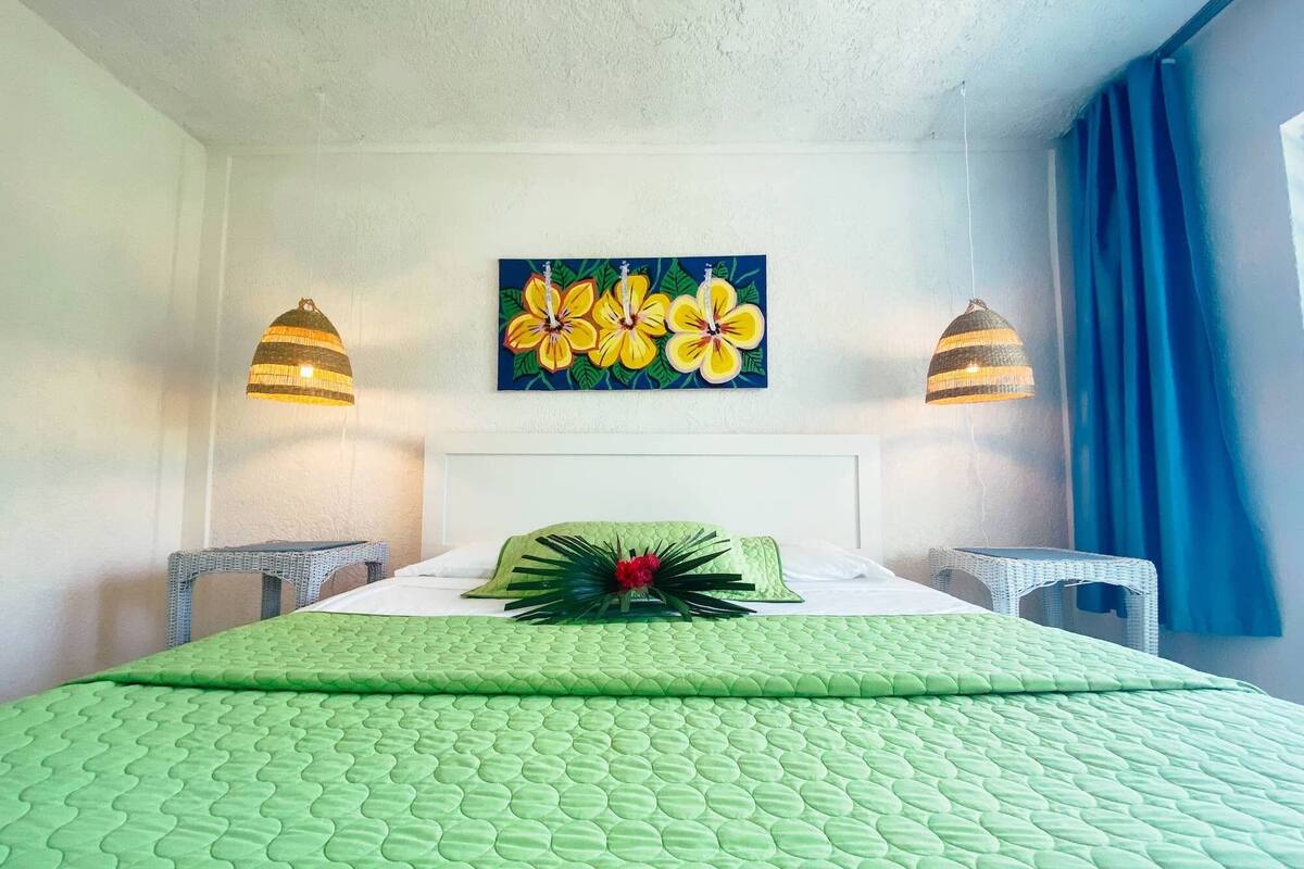 A hotel room with a green quilted bedspread, a floral painting above the bed, two wicker nightstands, and a blue curtain on the window.