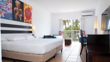 A hotel room with a large bed, a desk, a chair, and a balcony with a view of palm trees.
