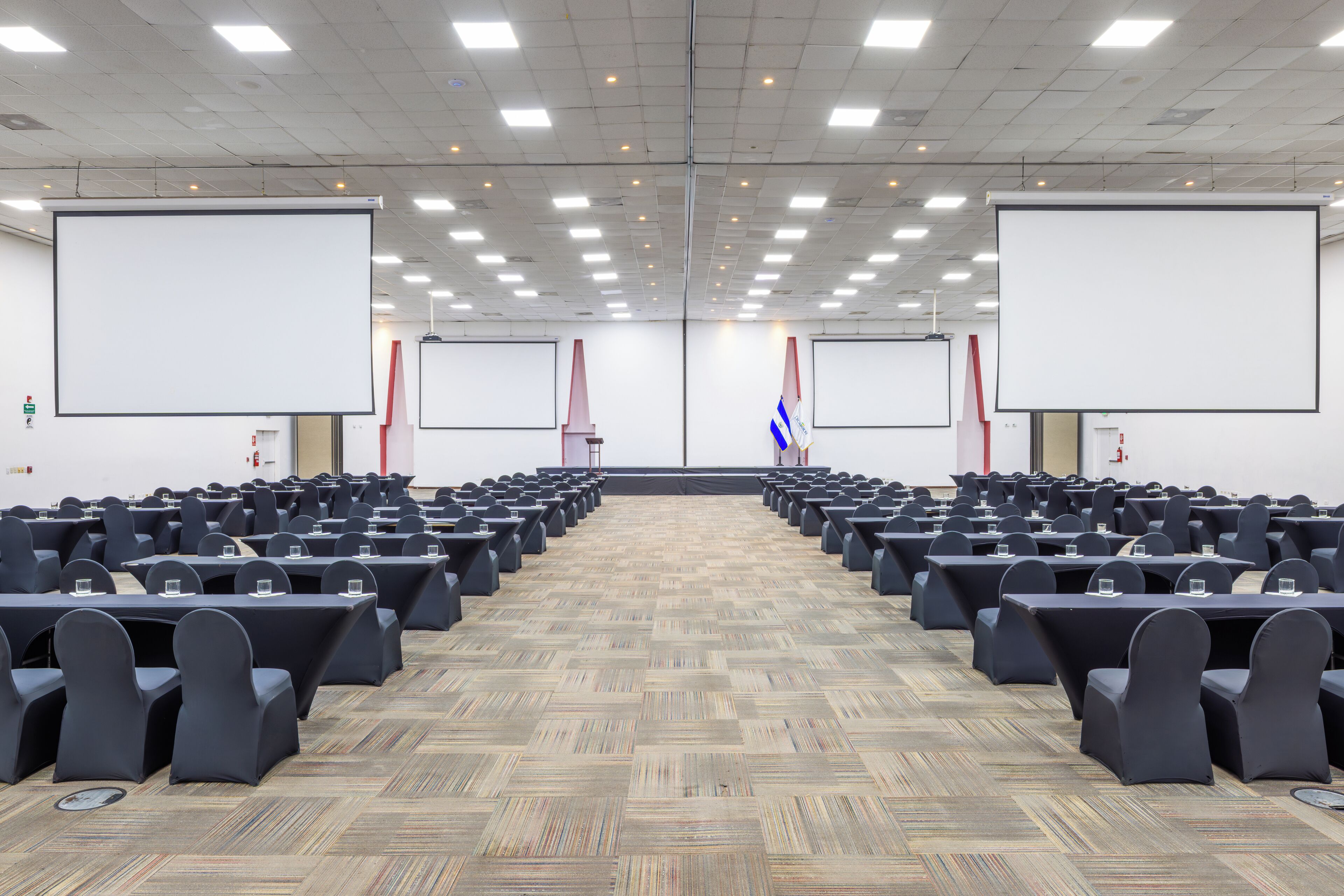 Une salle de conférence avec des rangées de tables et de chaises, de grands écrans de projection et un tapis à motifs.