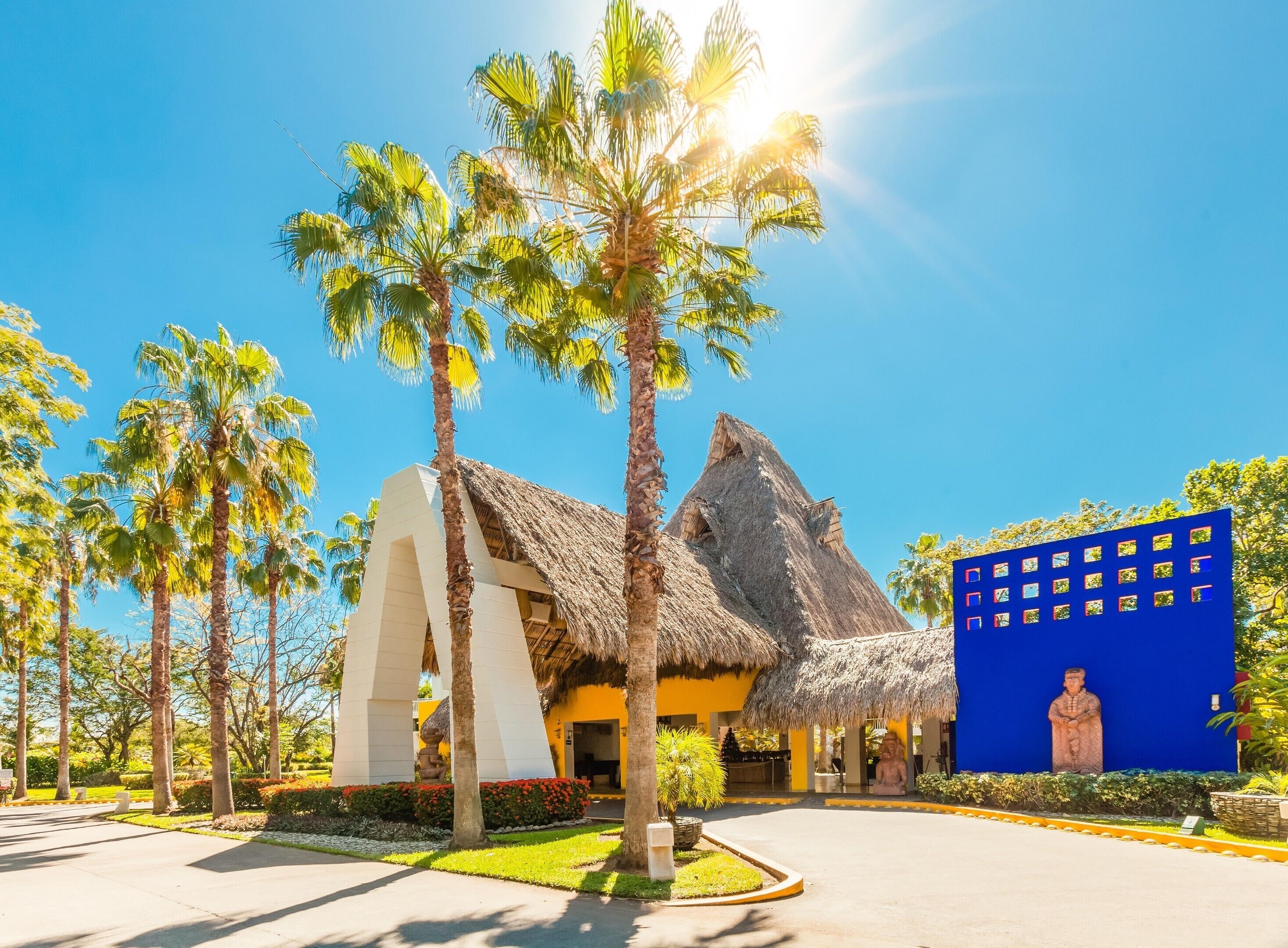 A resort with a thatched-roof building, palm trees, and a blue wall with a statue.