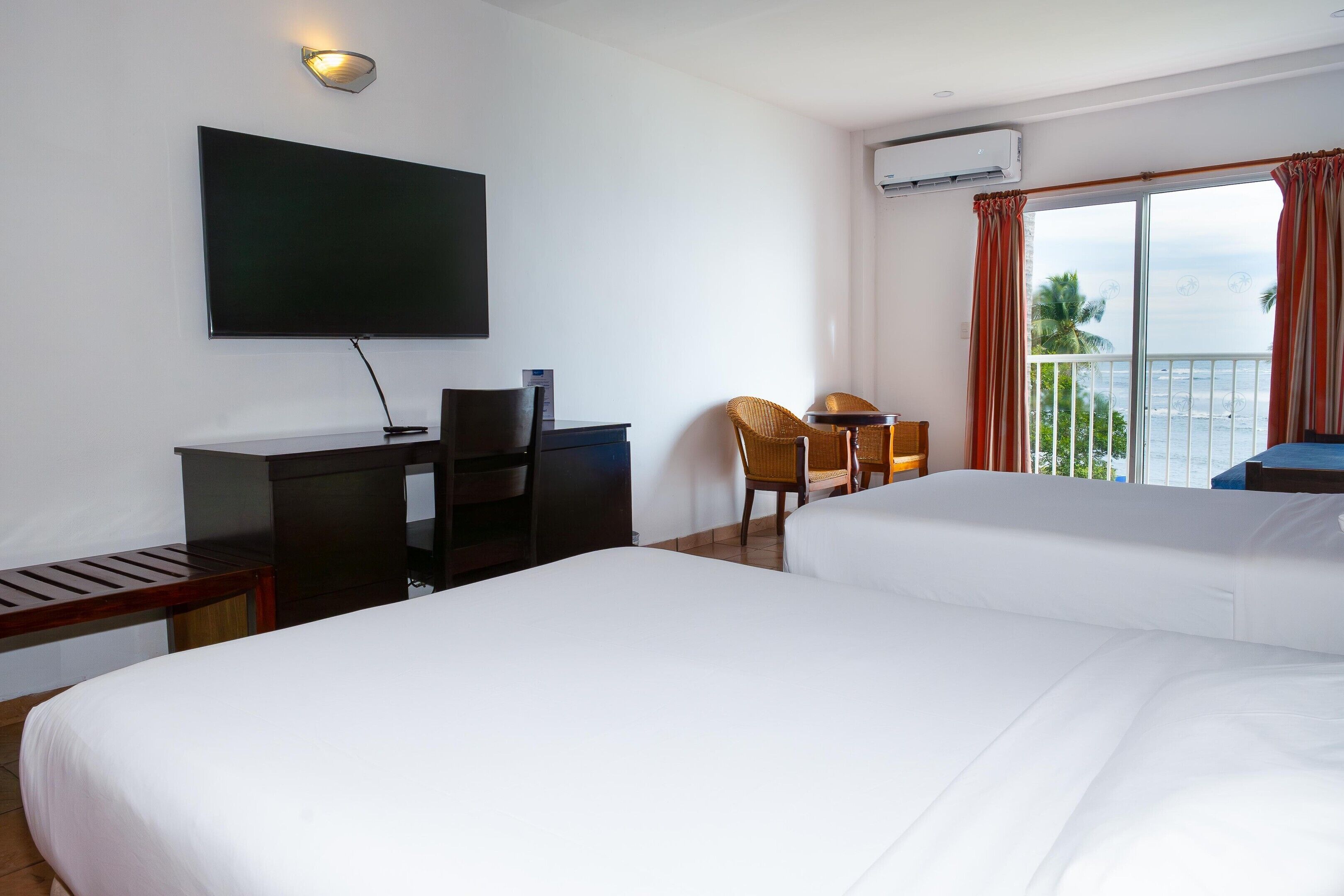 A hotel room with a bed, a desk, a chair, a TV, and a balcony with a view of the ocean.