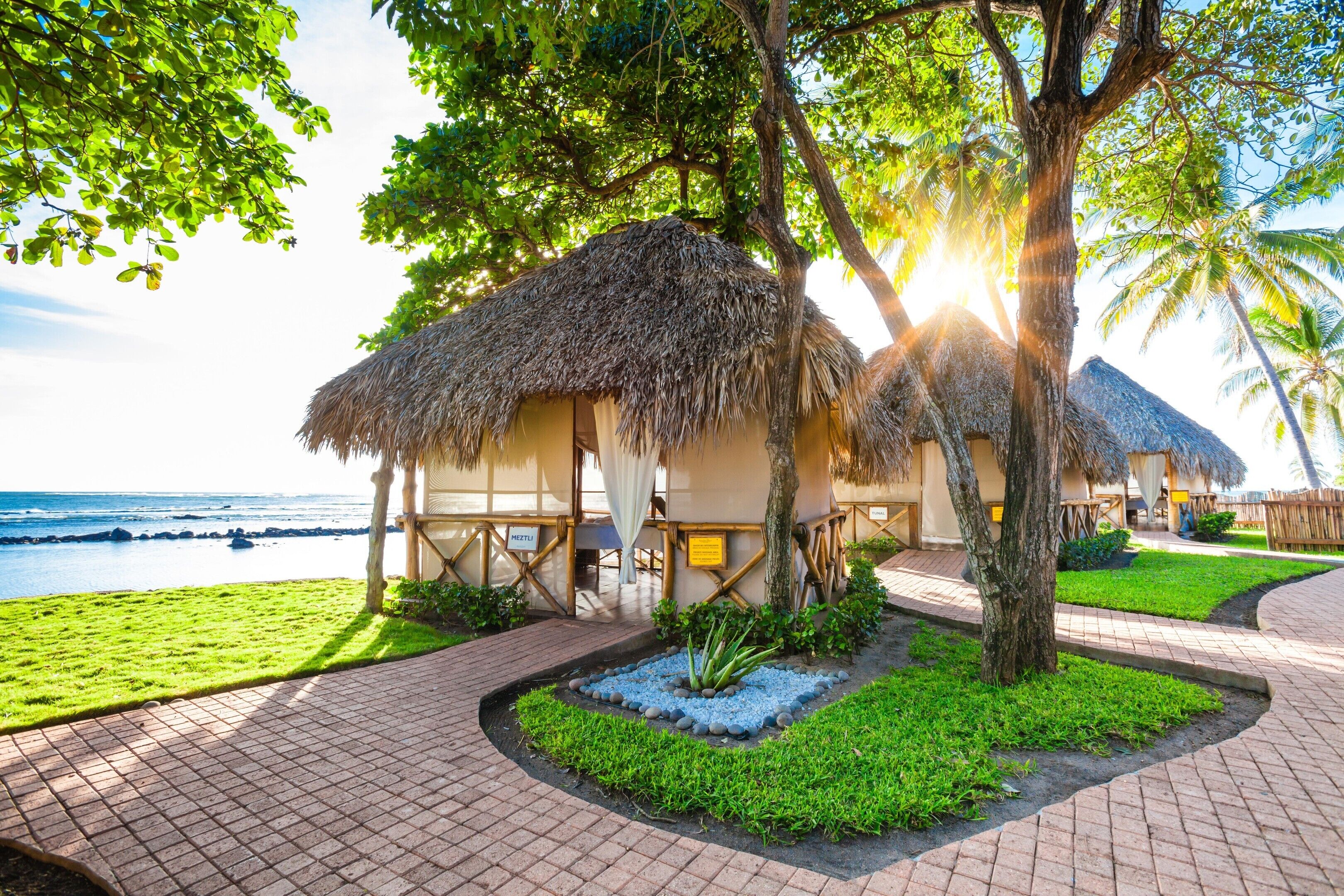 A beachfront resort with thatched-roof huts, a paved walkway, and a view of the ocean.