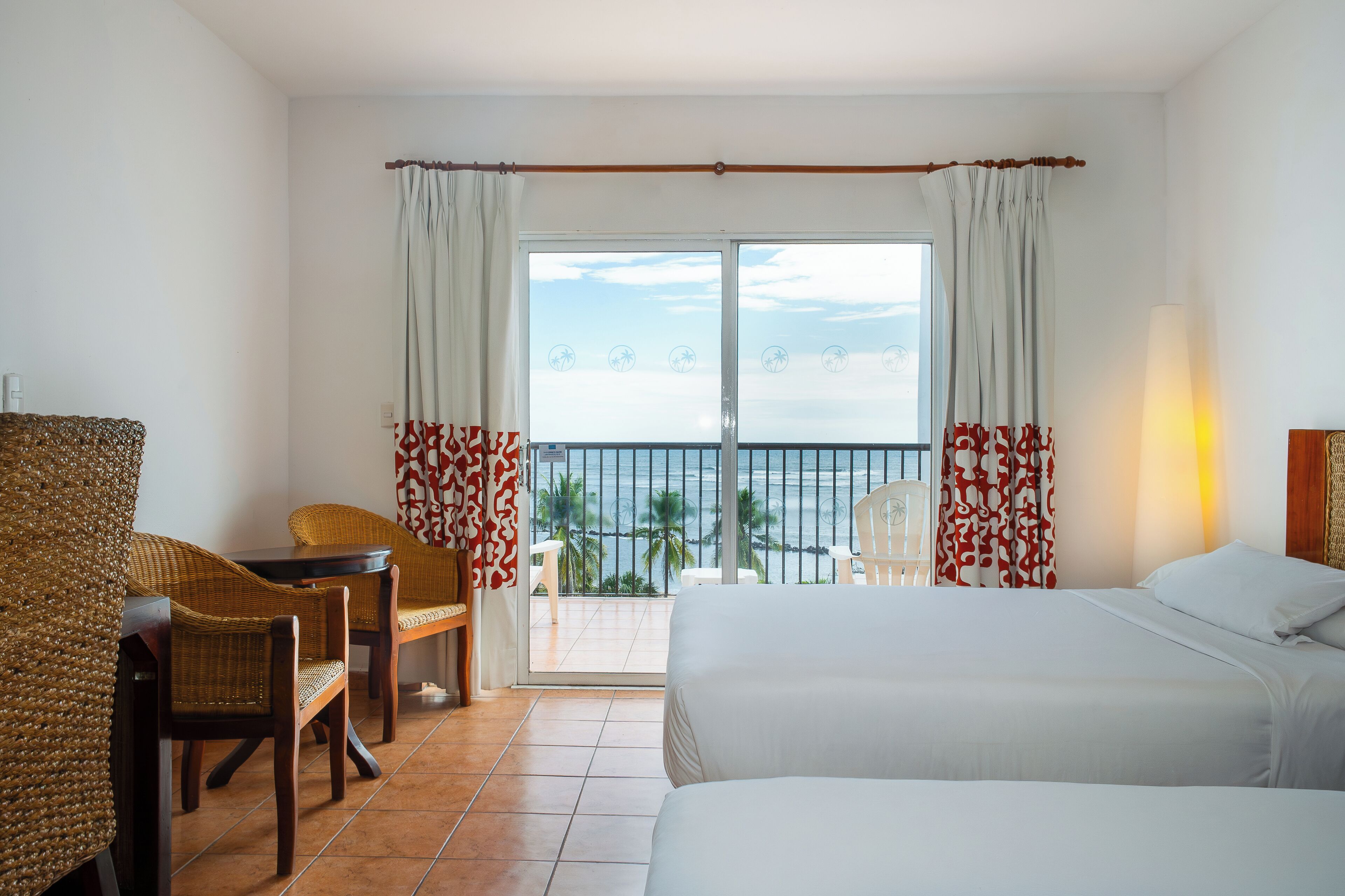 A hotel room with two beds, a dining area with wicker chairs, a table, and a view of the sea through a large window.
