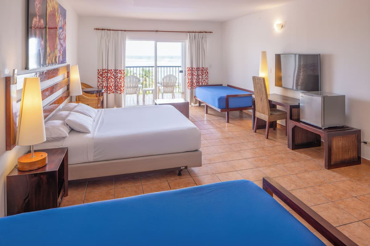 A hotel room with a bed, a desk with a television, a chair, and a view of the ocean through the balcony doors.