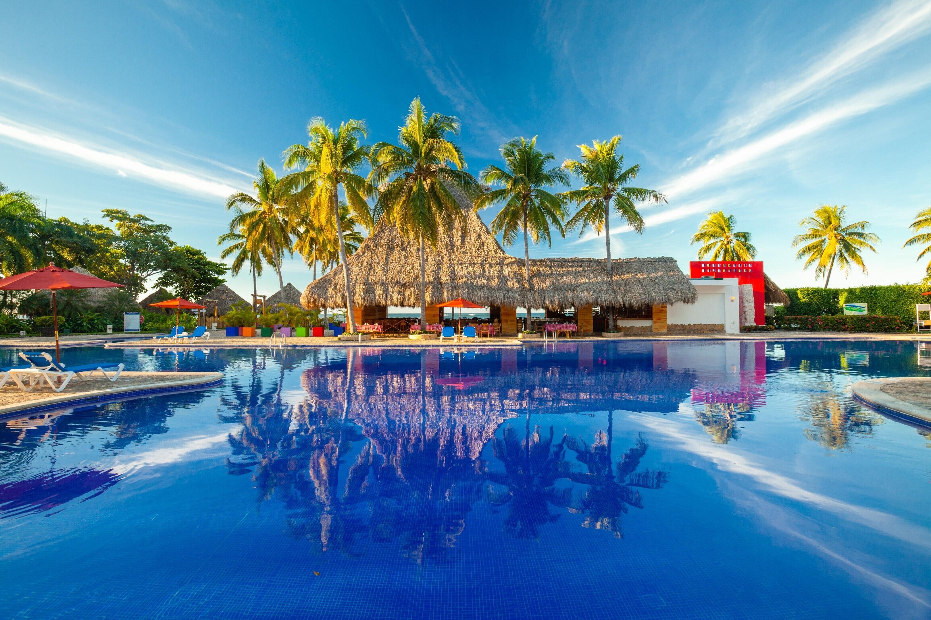 A pool with clear blue water reflecting palm trees and a thatched-roof building.