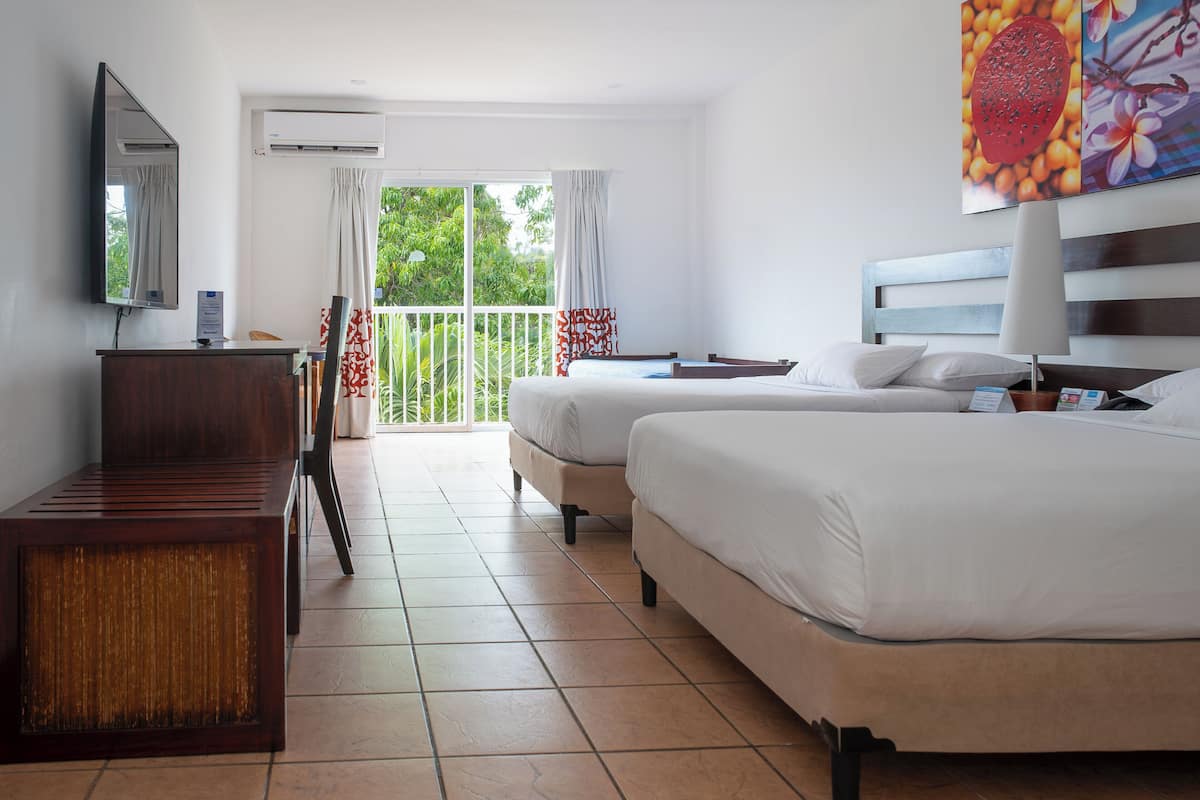 A hotel room with two beds, a desk, a TV, and a balcony with a view of greenery.