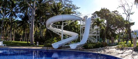 A swimming pool with a water slide, surrounded by palm trees and a clear sky.