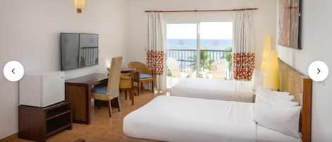A hotel room with two beds, a desk, a chair, a TV, and a balcony with a view of the sea.
