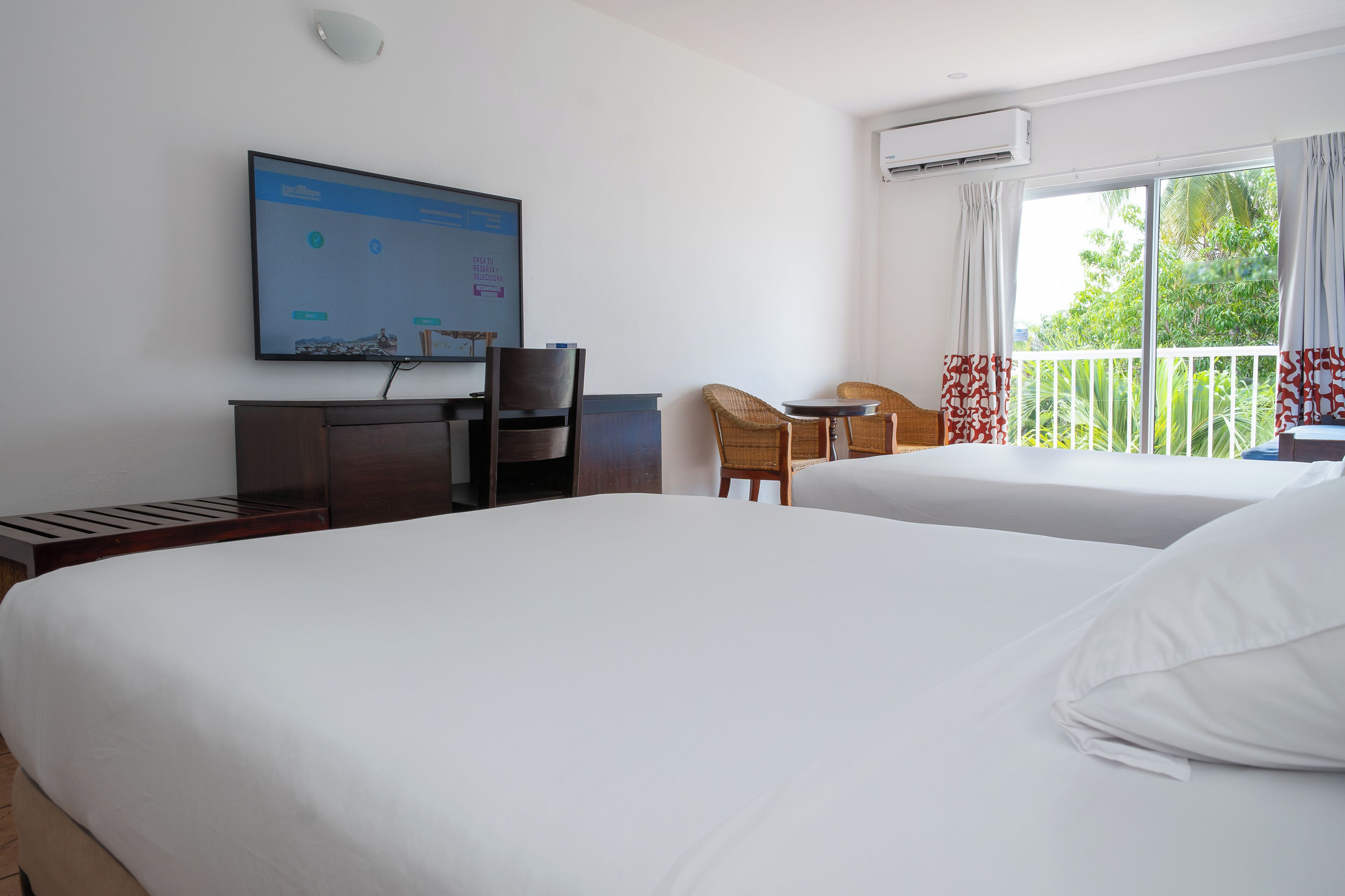 Ein Hotelzimmer mit zwei Betten, einem Schreibtisch, einem Stuhl, einem Fernseher und einem Balkon mit Vorhängen.