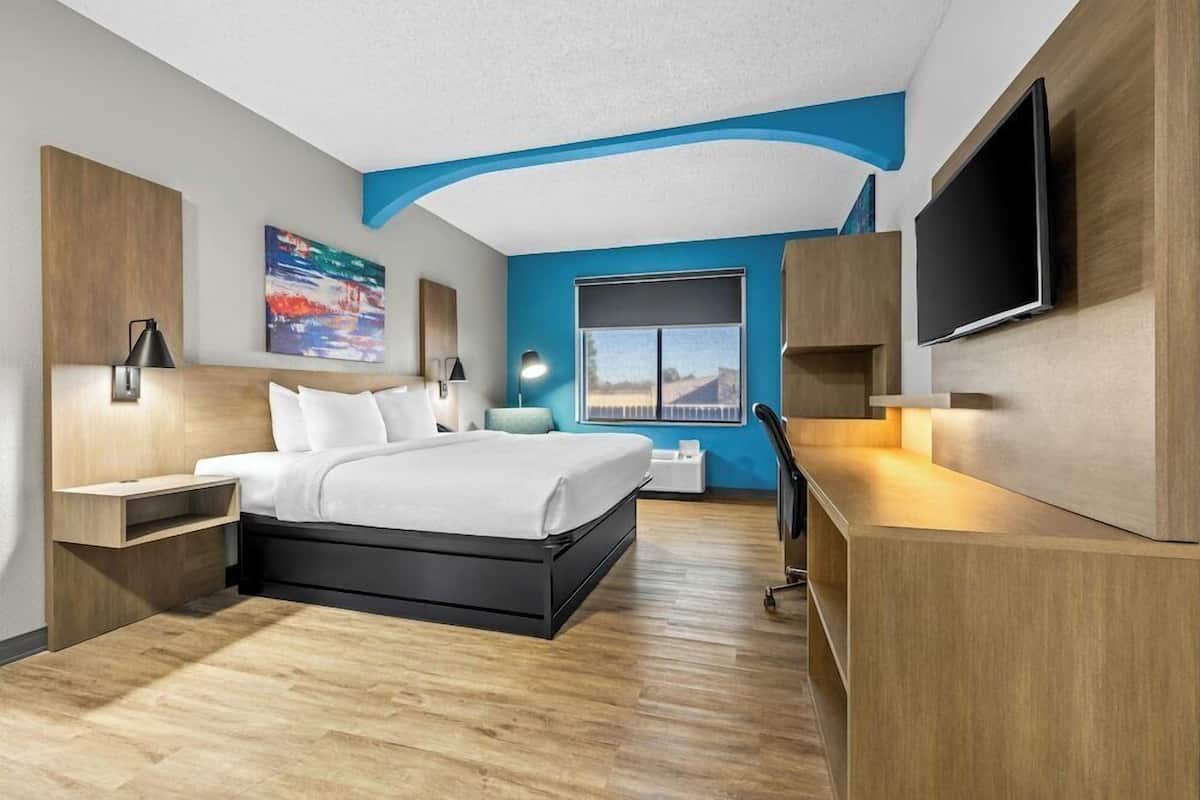 A modern hotel room with a bed, desk, TV, and artwork.