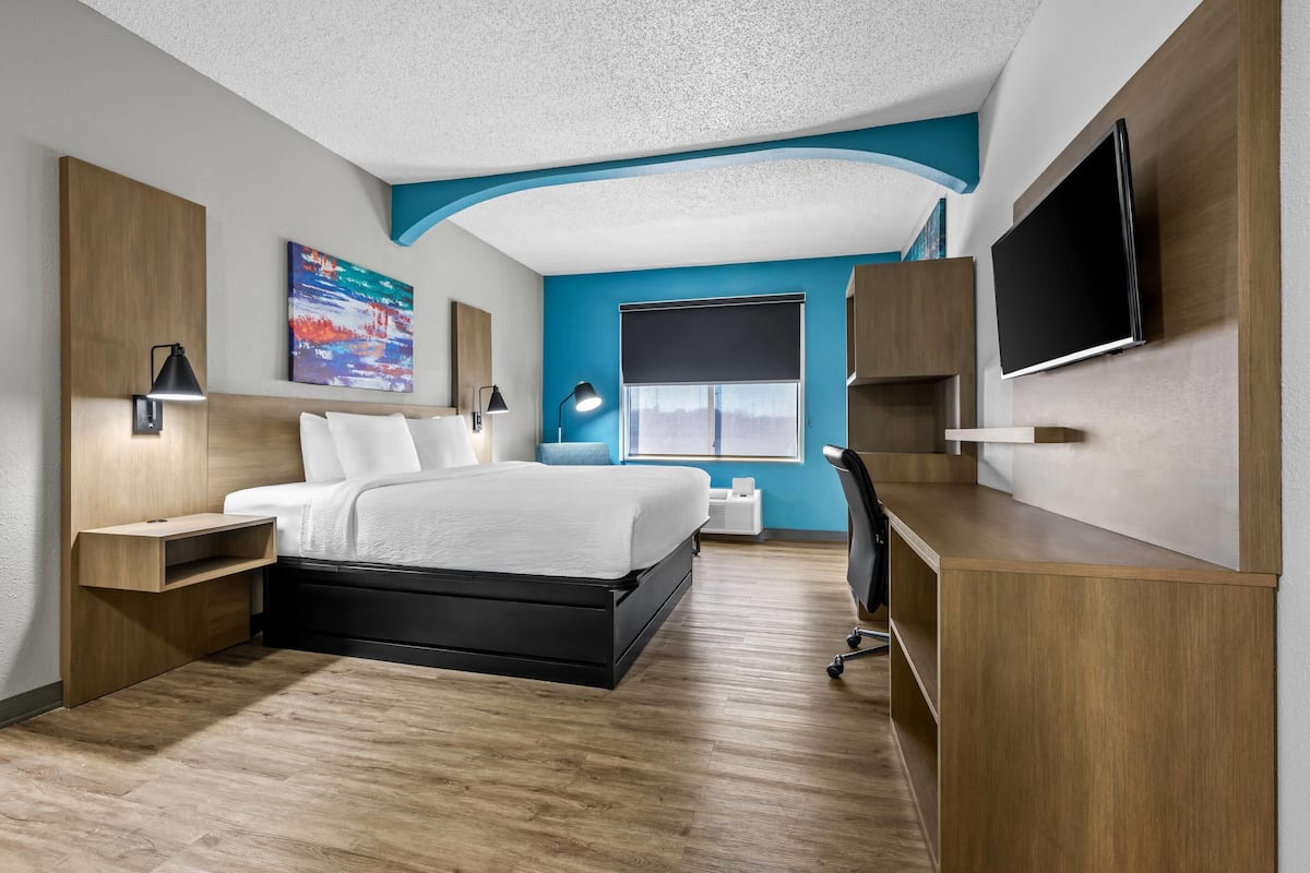A modern hotel room with a bed, desk, TV, and artwork.