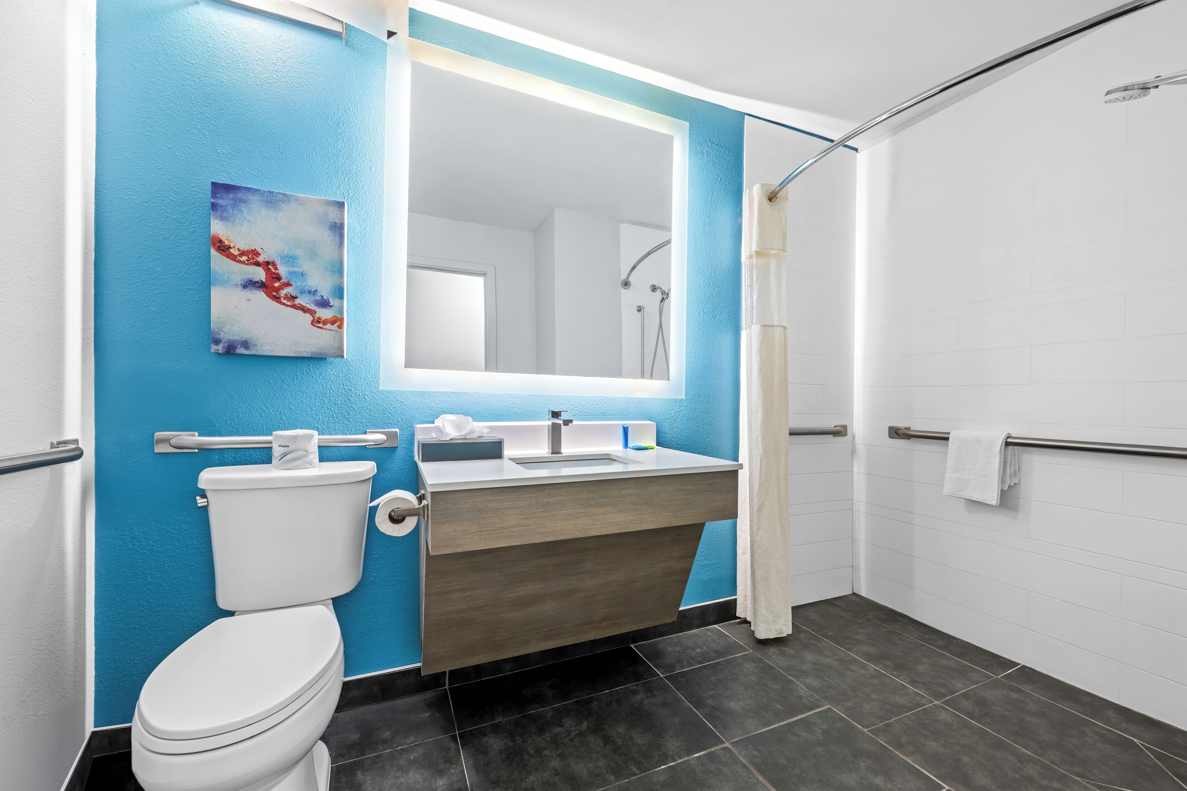 A modern bathroom with a blue accent wall, a white toilet, a wooden vanity, a large mirror, and a shower area.