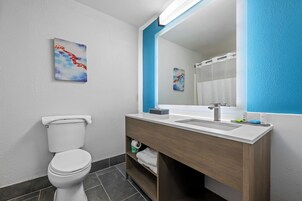 Un baño moderno con inodoro blanco, tocador de madera con lavamanos y un espejo grande. Hay dos obras de arte en las paredes.
