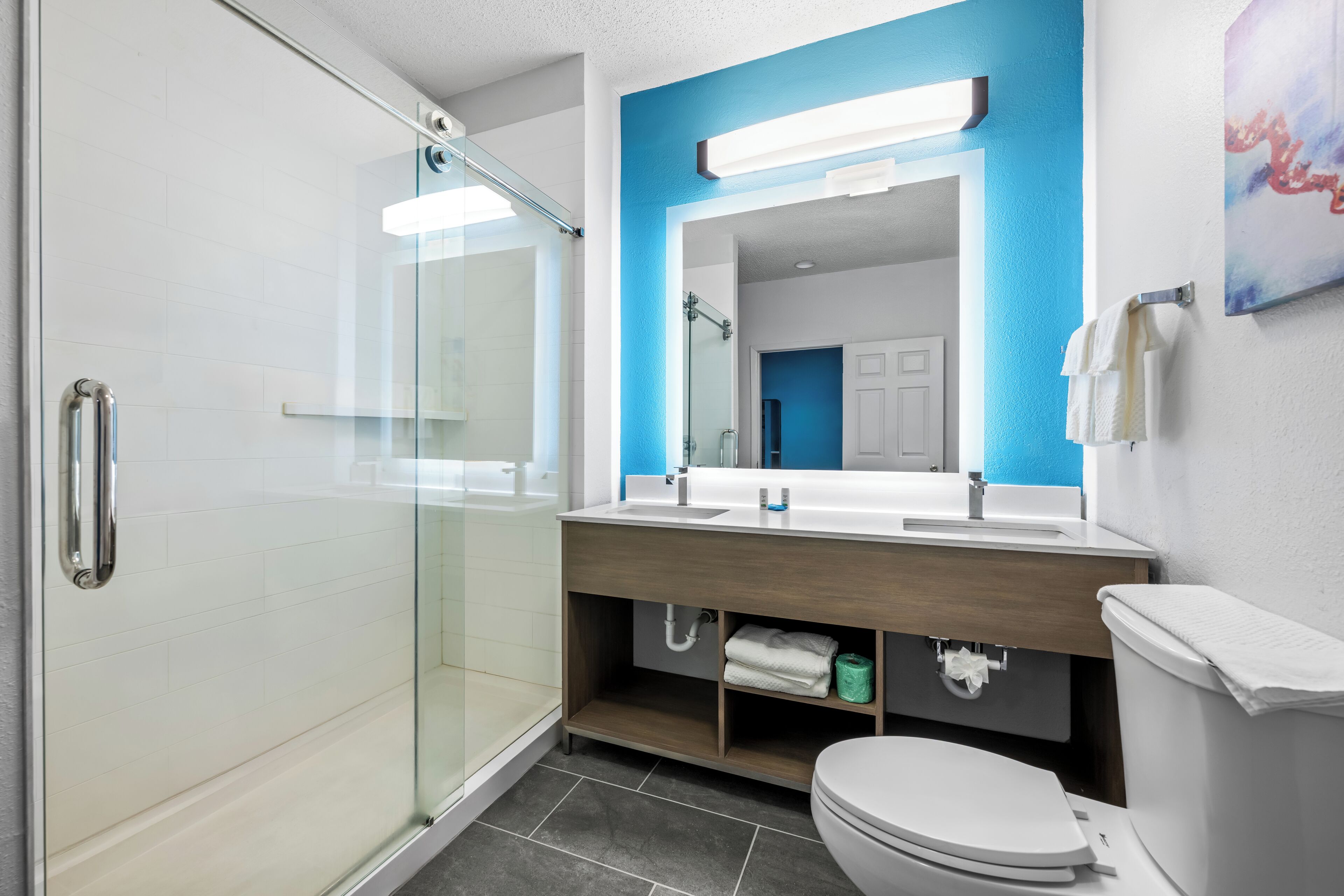 Un bagno moderno con box doccia in vetro, un water bianco, un mobile vanitario in legno con doppio lavabo e una parete d'accento blu.