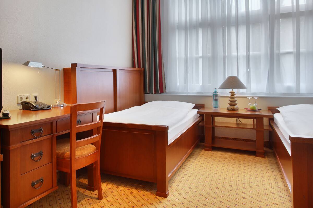 A hotel room with a wooden desk, a chair, a bed, and a bedside table with a lamp and a bottle.
