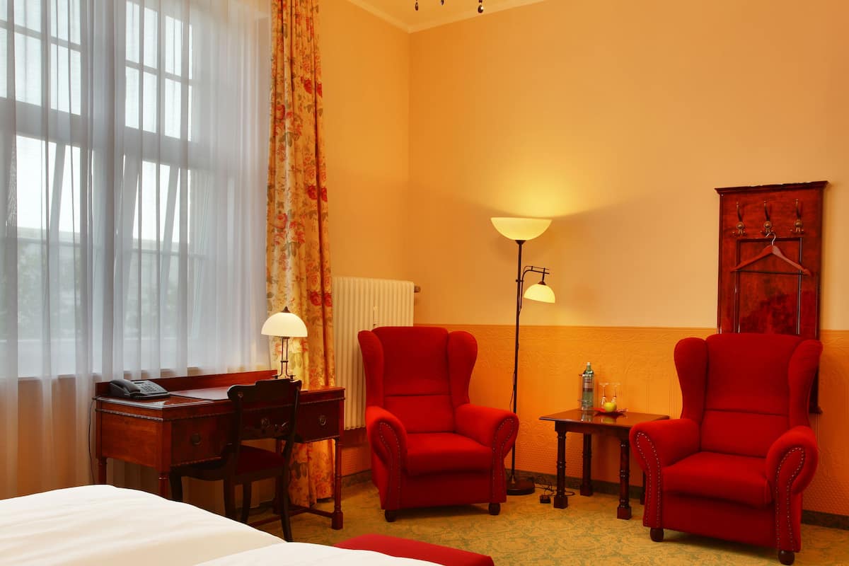 A hotel room with a bed, two red armchairs, a desk with a lamp, a radiator, and a wall-mounted coat rack.