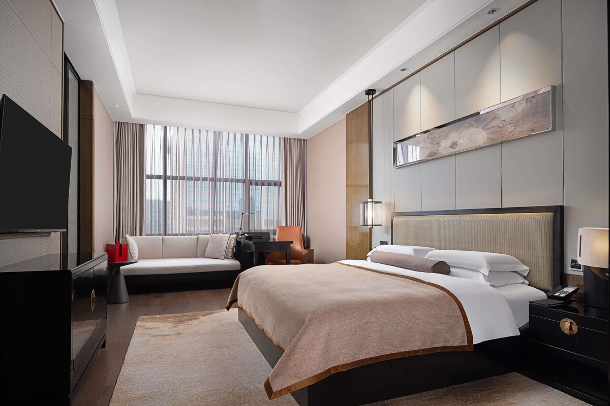 A modern hotel room with a large bed, a TV, a desk, and a window with curtains.