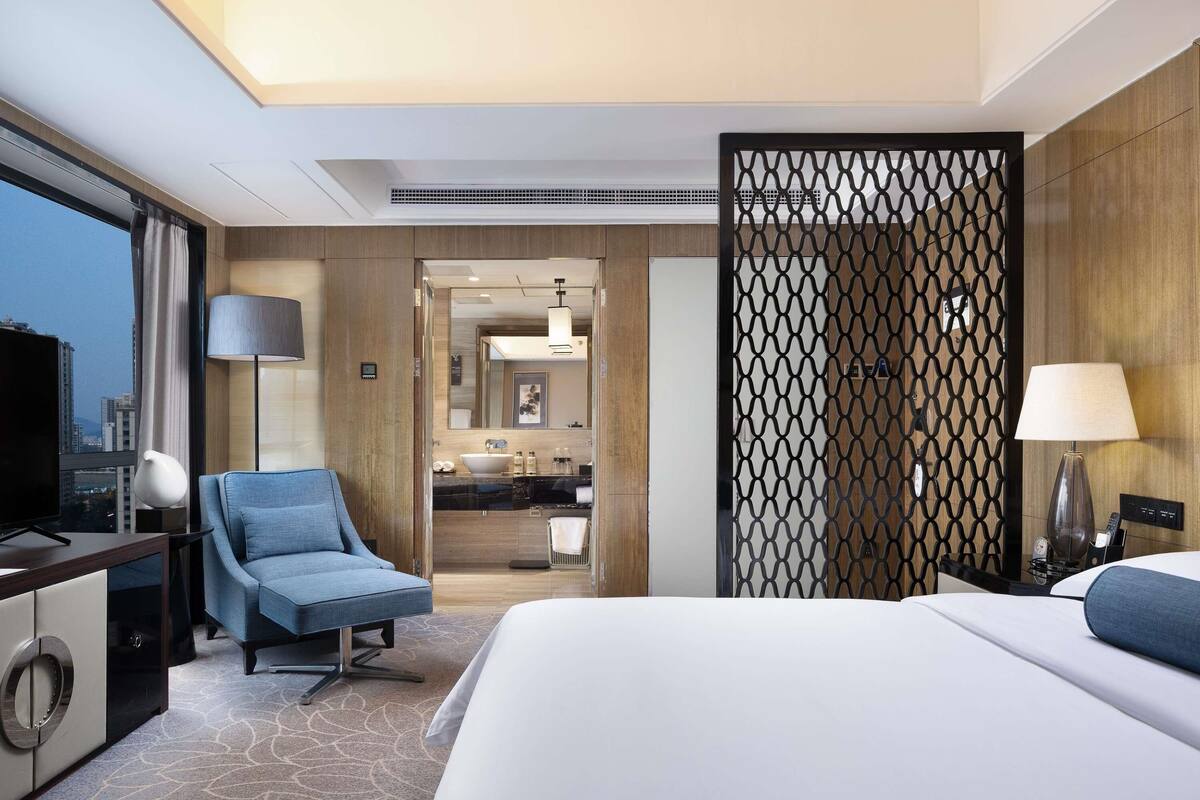 Habitación de hotel con una cama grande, zona de estar con una silla, escritorio con televisor, baño con lavabo y espejo, y vistas a la ciudad.