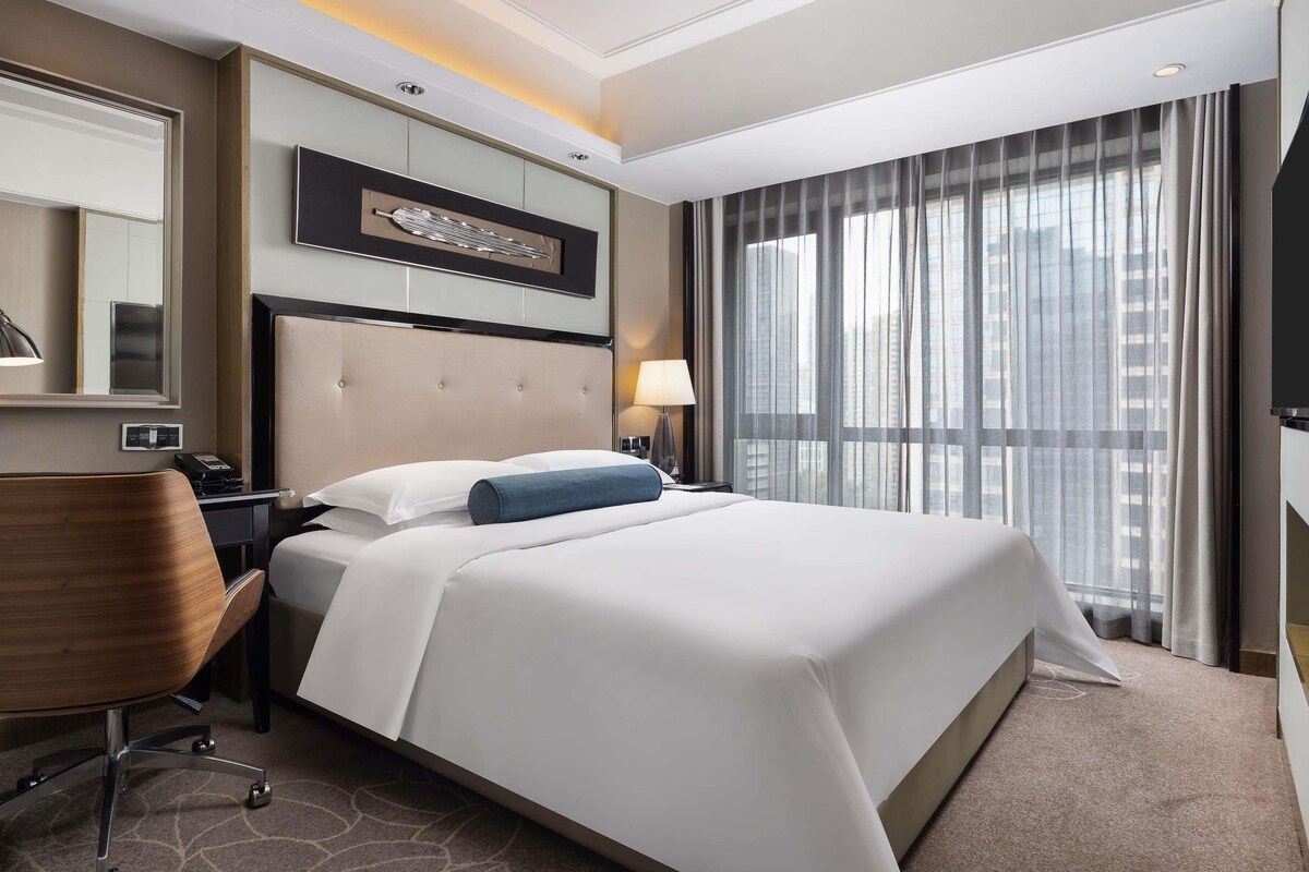 A hotel room with a large bed, a desk, a chair, a mirror, and a window with curtains.