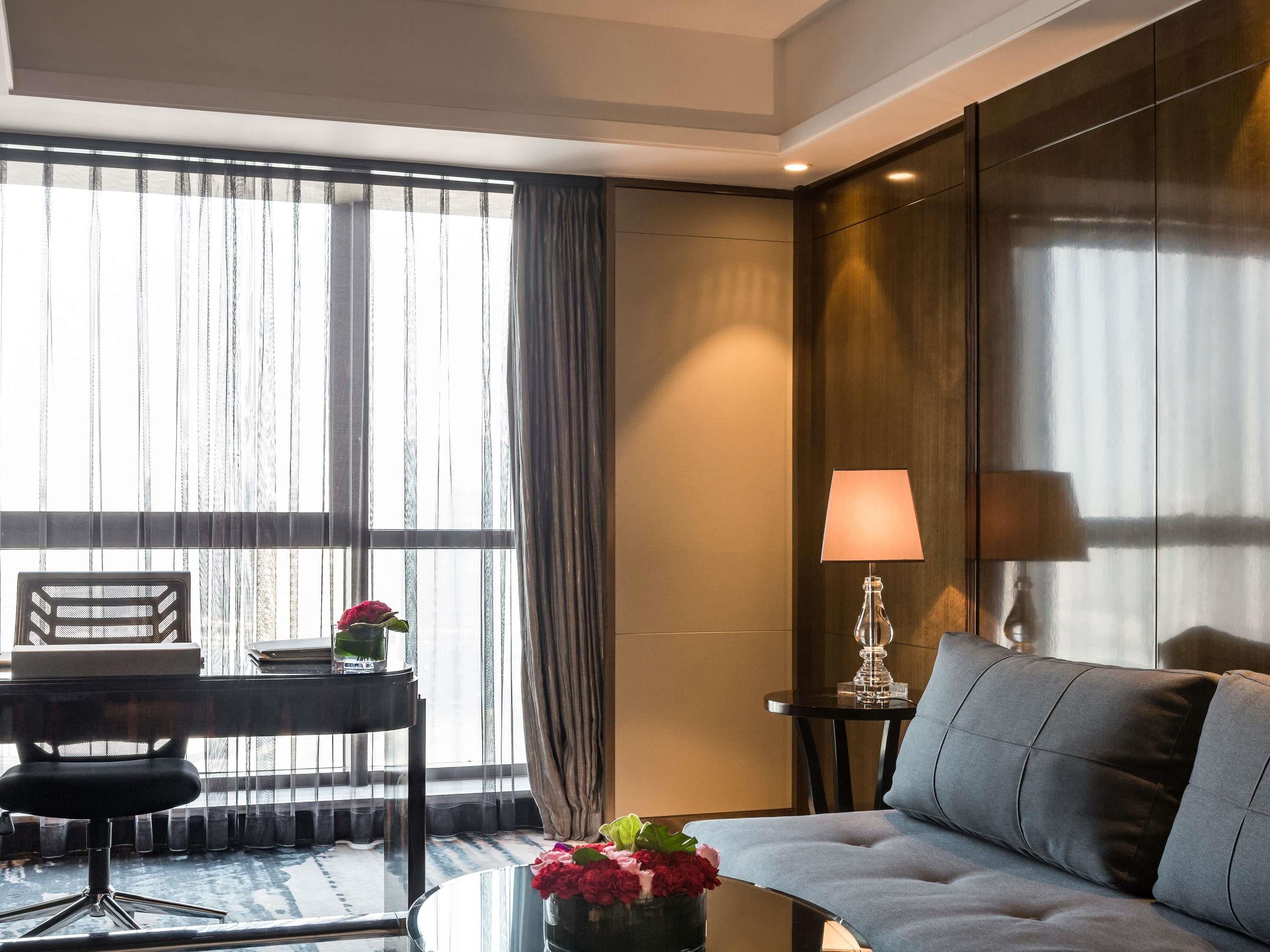 A modern hotel room with a sofa, a desk with a chair, a lamp, and a vase with flowers.