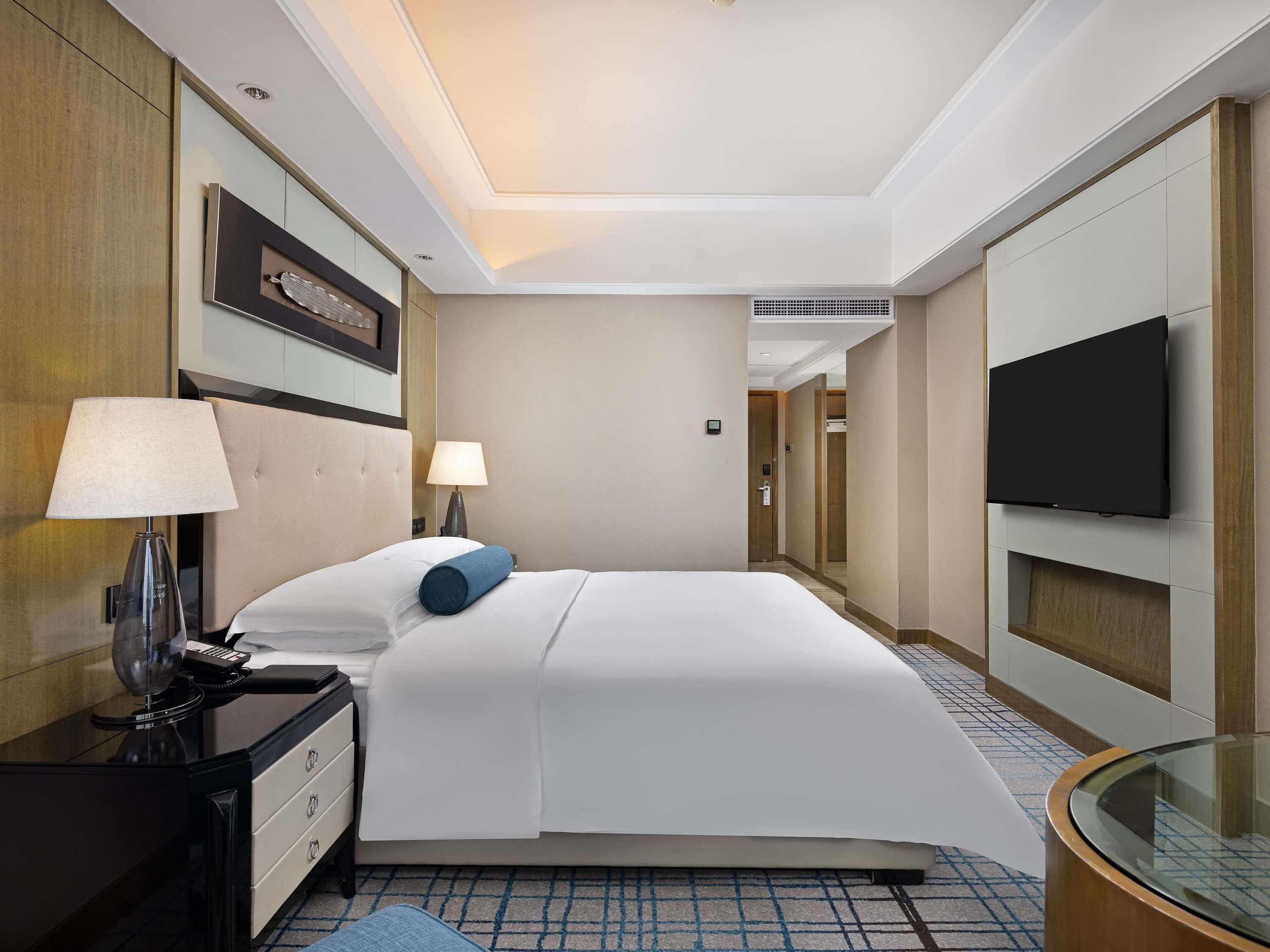 A hotel room with a bed, bedside lamps, a desk with a telephone, a television mounted on the wall, and a glass table.