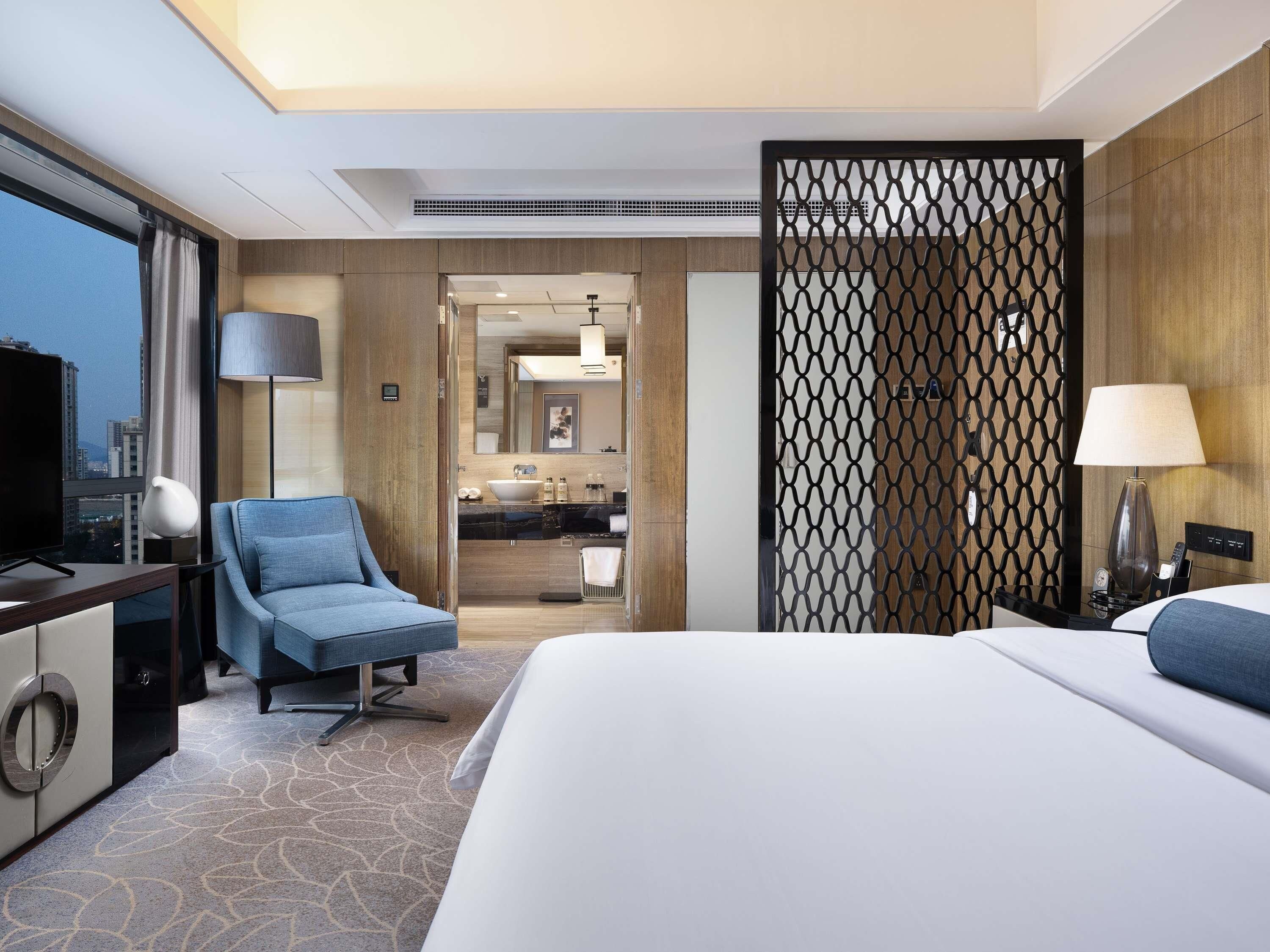 A hotel room with a large bed, a seating area with a chair, a desk with a TV, a bathroom with a sink and mirror, and a view of the city.