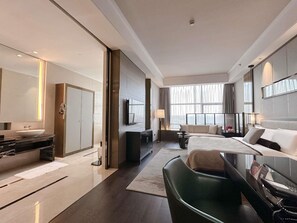 Quarto de hotel moderno com cama grande, sofá, escrivaninha, televisão e banheiro com pia e espelho.