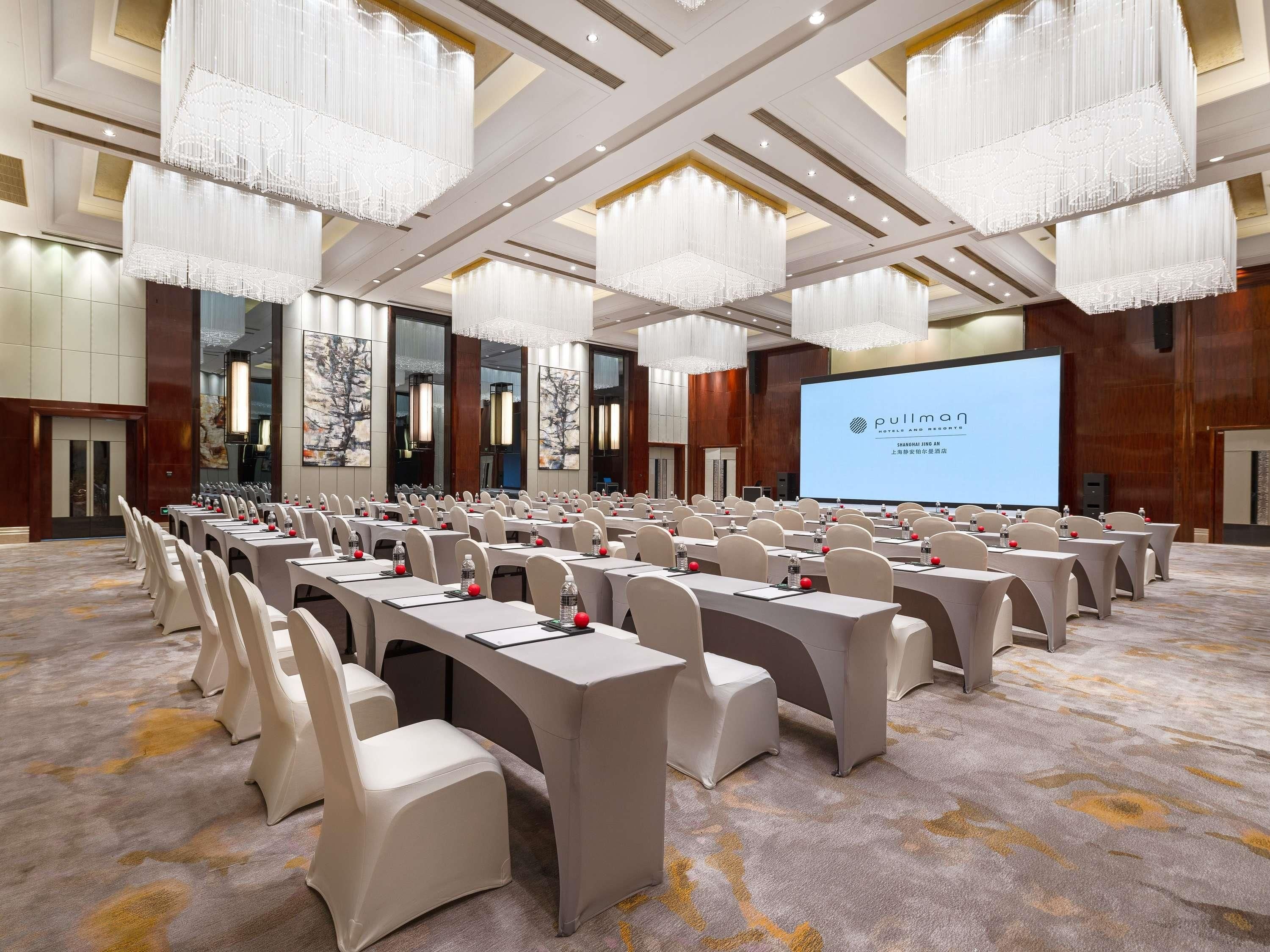 A conference room with rows of tables and chairs, a large screen, and chandeliers.