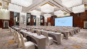 A conference room with rows of tables and chairs, a large screen, and chandeliers.