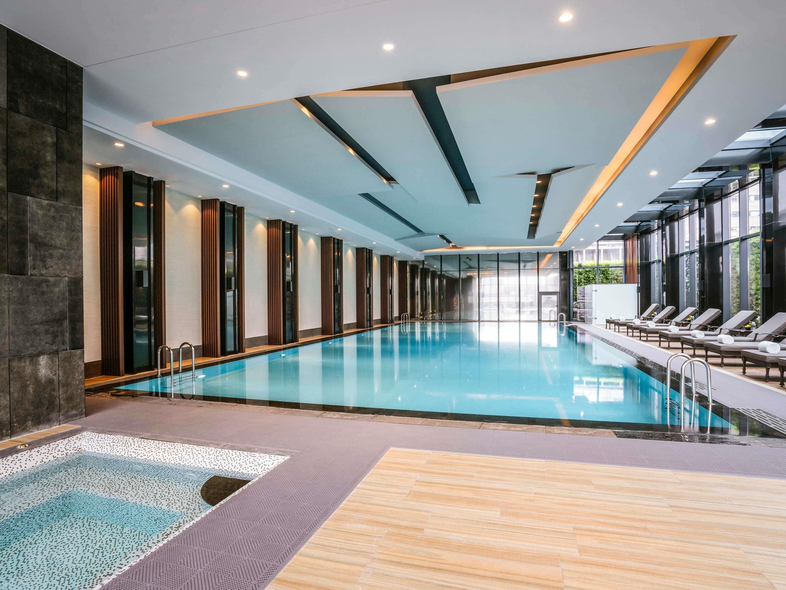 Una piscina interior moderna con un diseño largo y esbelto, rodeada de sillones de descanso y un muro de cristal.