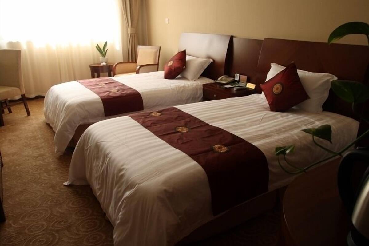 Quarto de hotel com duas camas, uma mesinha, uma cadeira e um vaso de planta.