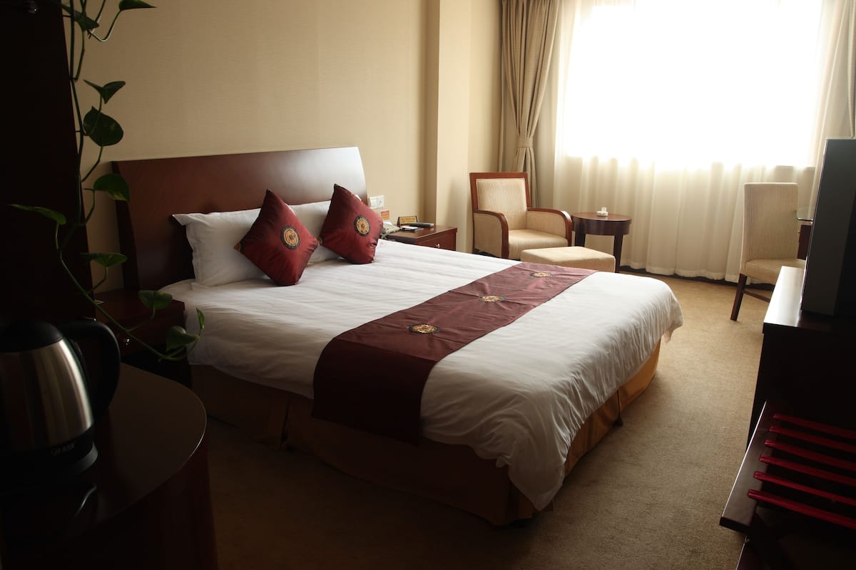 Quarto de hotel com uma cama grande, duas cadeiras, uma mesinha e uma televisão.
