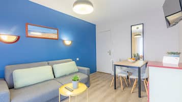Een moderne woonkamer met een blauwe accentmuur, een grijze bank, een klein rond tafeltje met een plant, en een eethoek met een houten tafel en stoelen.