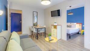 Habitació d'hotel moderna amb sofà, taula de menjador, cadires, televisió, llit i taula petita amb comandament a distància.