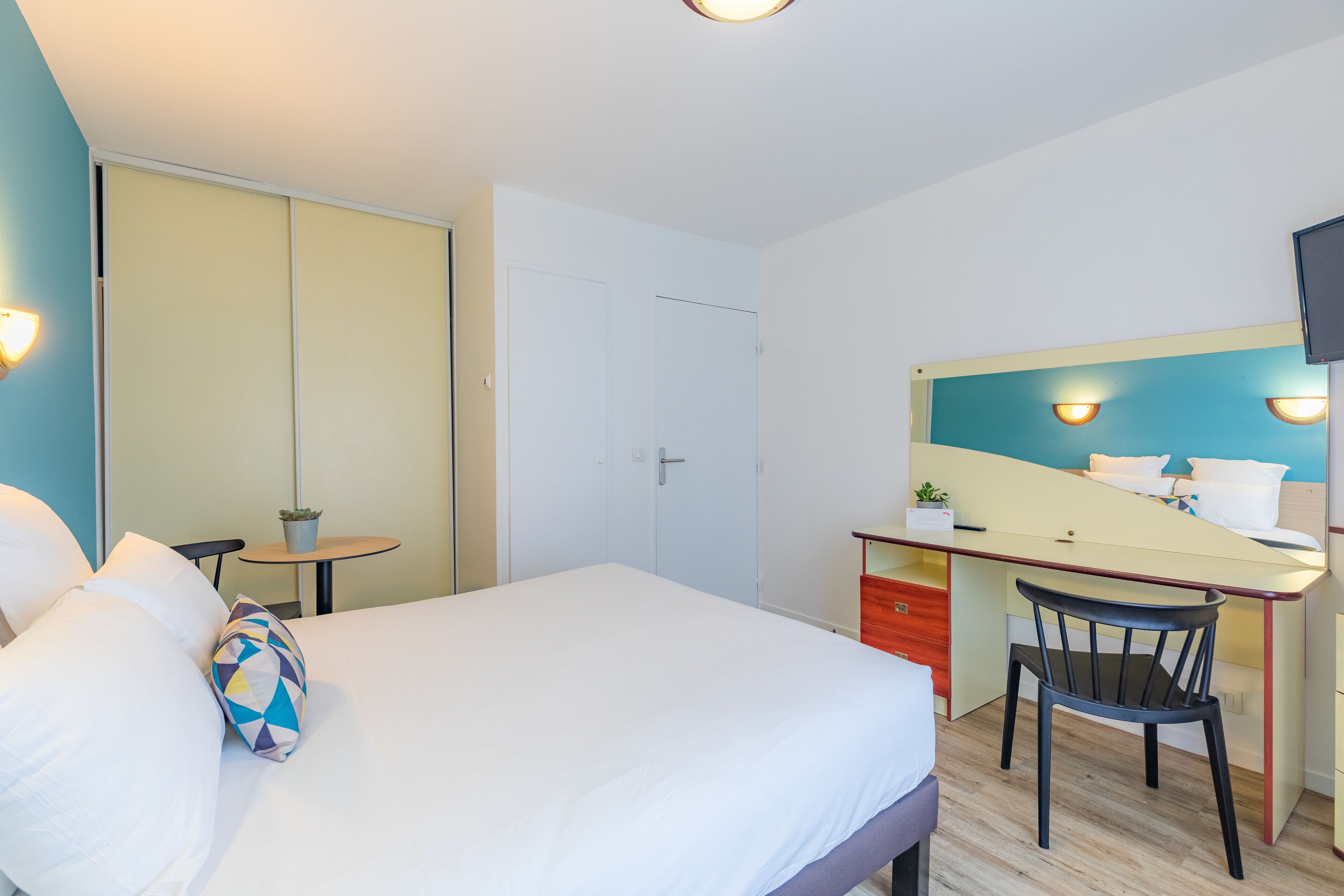 A hotel room with a bed, a desk with a chair, a small table with a plant, and a TV mounted on the wall.