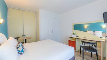 A hotel room with a bed, a desk with a chair, a small table with a plant, and a TV mounted on the wall.
