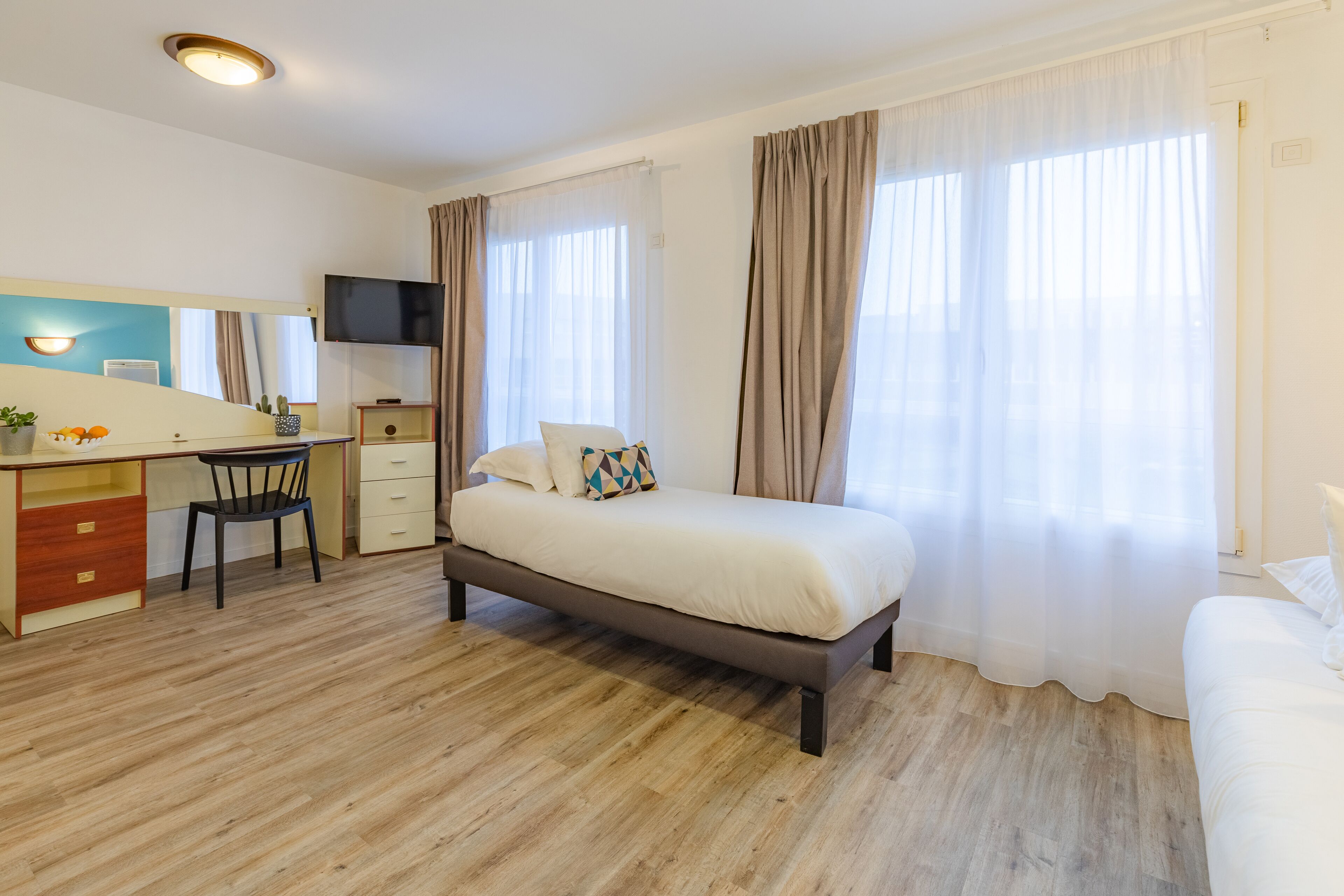 Une chambre d’hôtel équipée d’un lit, d’un bureau, d’une chaise, d’une télévision et de grandes fenêtres avec des rideaux.